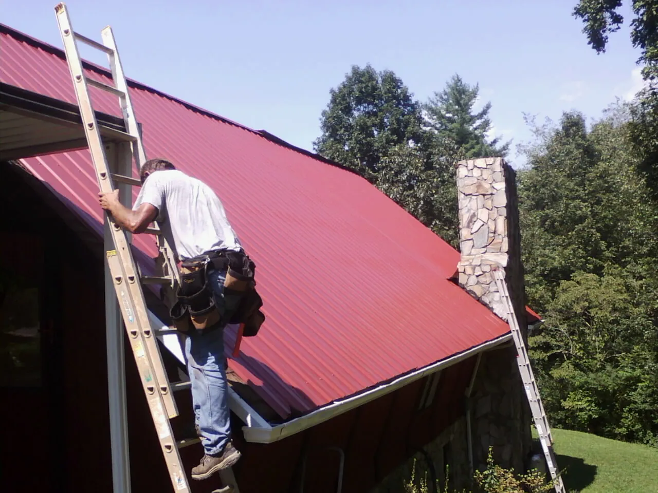Roofer on a ladder beside a red metal roof and stone chimney — Soffit & Fascia Repair by Roofing Brothers in Spencerville